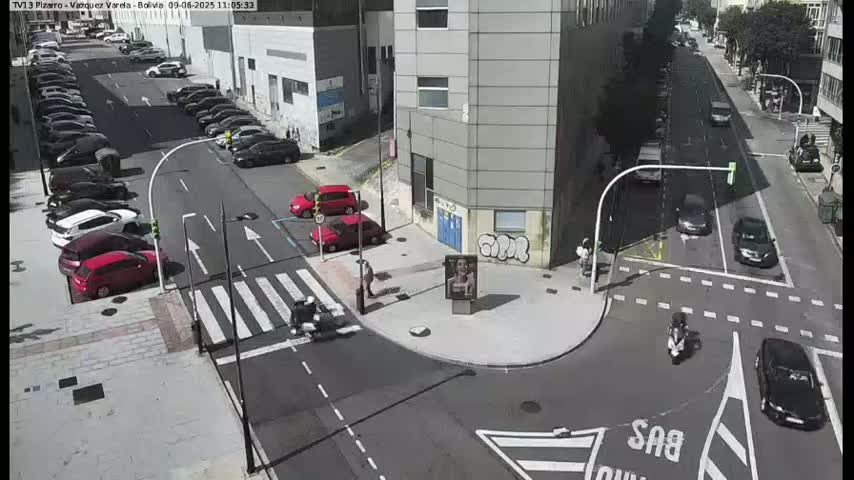 A sunny city intersection shows several cars parked along the street and others driving through a crosswalk, with a few motorcyclists present.