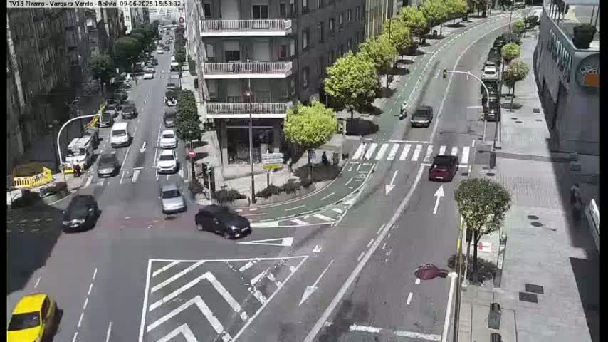 A sunny city street intersection shows several cars and a pedestrian crossing, with bike lanes and a building in the background.