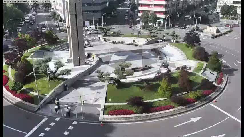 An aerial view shows a circular plaza with landscaped greenery, walkways, and a modern building, on a sunny day.