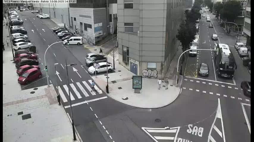 A street intersection with parked and moving cars, a pedestrian crossing, and buildings on a partly cloudy day.