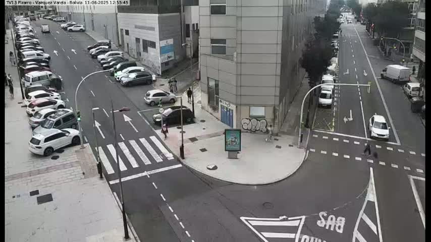 A city street intersection with parked cars lining the sides, several cars in motion, and pedestrians crossing at crosswalks, on a mostly clear day.