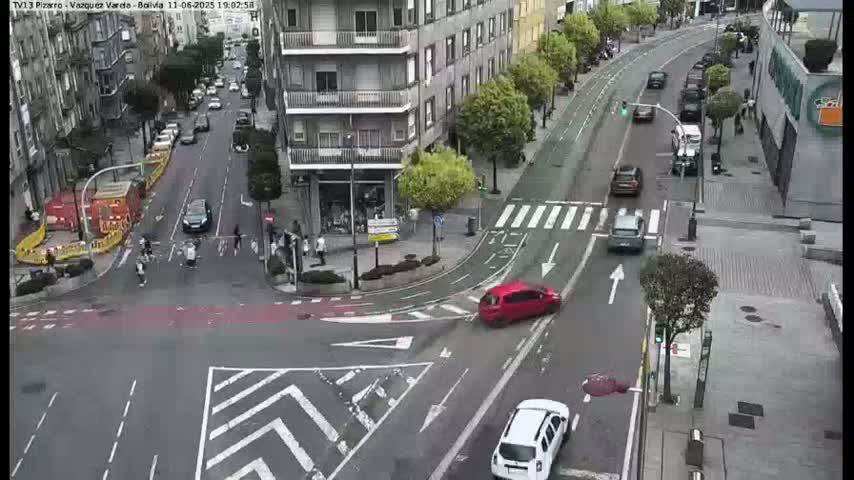 A street intersection with several cars and pedestrians, under a mostly clear sky.