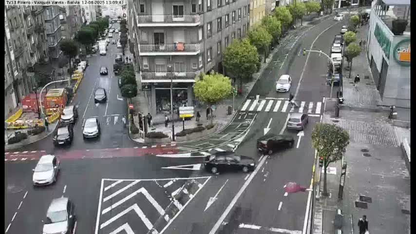 A wet city street intersection shows several cars navigating a curved road, with buildings and pedestrians visible in the background.