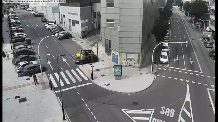 A sunny city street scene shows a corner intersection with parked cars along the sides of the street and a few cars driving through.
