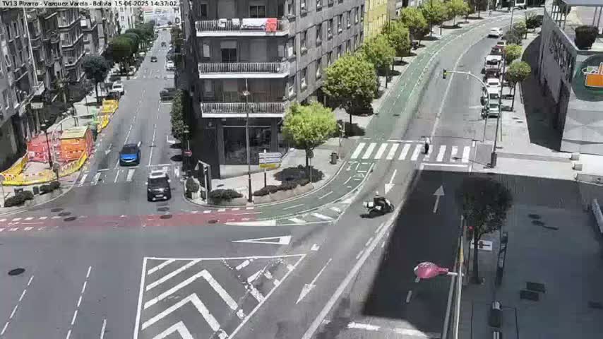 A high-angle view of a city intersection on a sunny day, with several cars and a motorcycle at a crosswalk, alongside buildings and a bike lane.