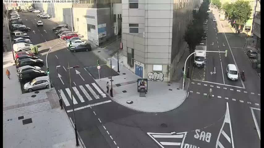 A street intersection with parked cars and a few pedestrians, under a sunny sky.