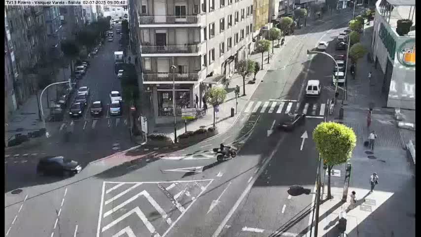 A sunny day reveals a city street intersection with several cars and pedestrians, a motorcyclist, and buildings lining the streets.