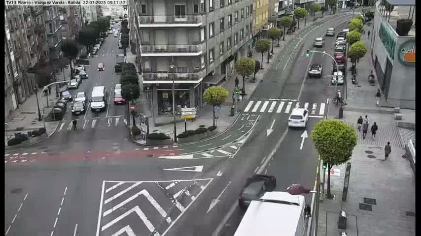 A city street intersection with several cars and pedestrians, under overcast skies.