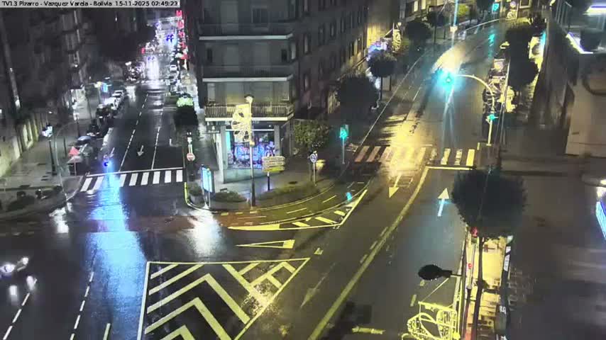 An elevated view captures a wet city street at night, with bright reflections shimmering on the rain-slicked pavement from streetlights and traffic signals, surrounded by buildings and a few visible vehicles under rainy conditions.