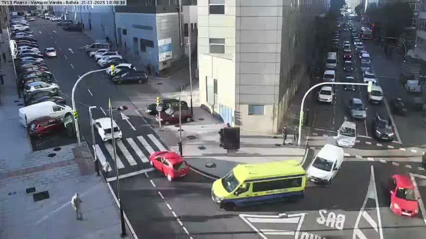 An overhead view captures a busy urban intersection on a clear and sunny day, with cars moving and stopped, including a prominent yellow and blue medical van and a red car in the foreground, alongside parked vehicles and pedestrians.
