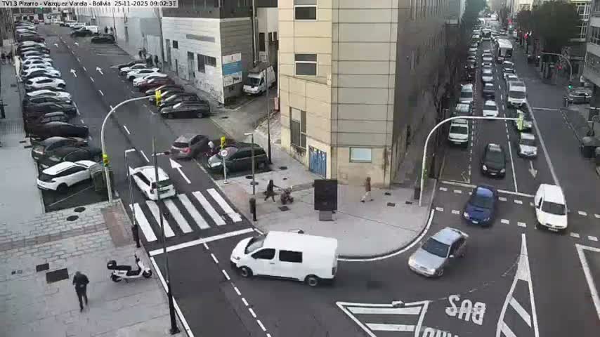 An overhead view captures a bustling city street intersection with parked cars, vehicles moving through a crosswalk, heavy traffic queuing down one street, and several pedestrians on sidewalks, all under overcast skies.