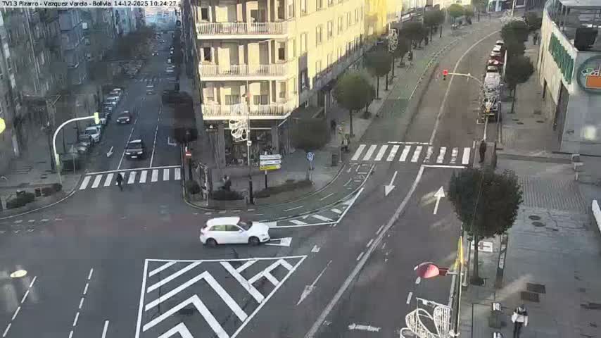 An overhead view captures a bustling city street intersection with parked cars, vehicles moving through a crosswalk, heavy traffic queuing down one street, and several pedestrians on sidewalks, all under overcast skies.