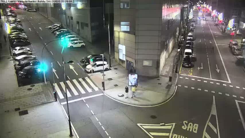 An overhead view captures a bustling city street intersection with parked cars, vehicles moving through a crosswalk, heavy traffic queuing down one street, and several pedestrians on sidewalks, all under overcast skies.