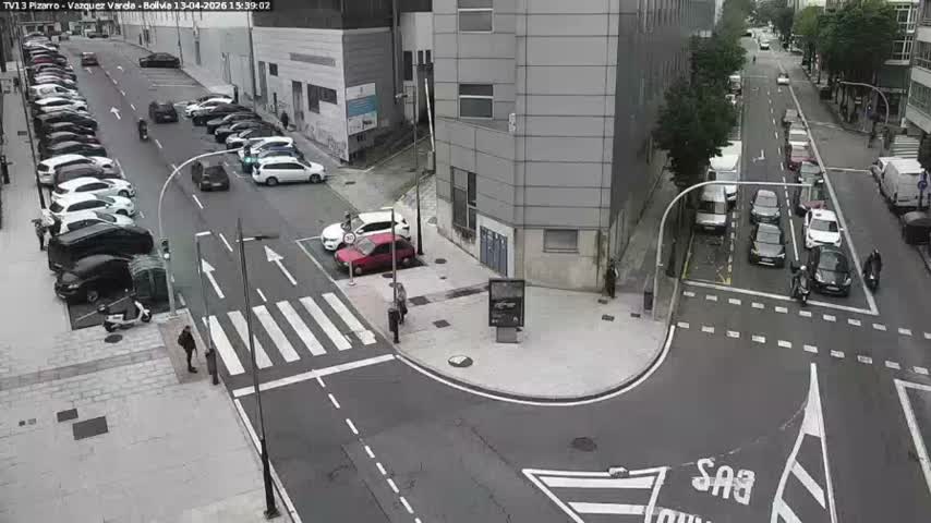 An overhead view captures a bustling city street intersection with parked cars, vehicles moving through a crosswalk, heavy traffic queuing down one street, and several pedestrians on sidewalks, all under overcast skies.