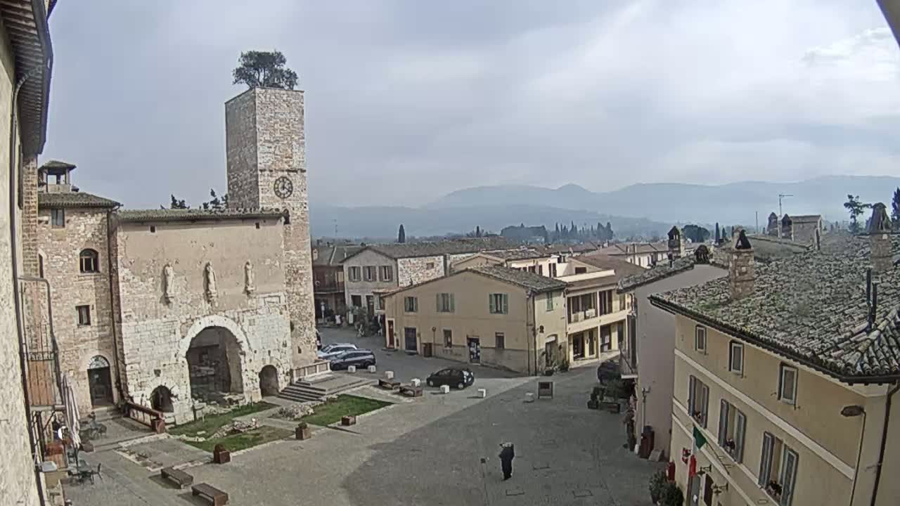 A town square with stone buildings, a clock tower topped with a tree, and distant mountains under an overcast sky.