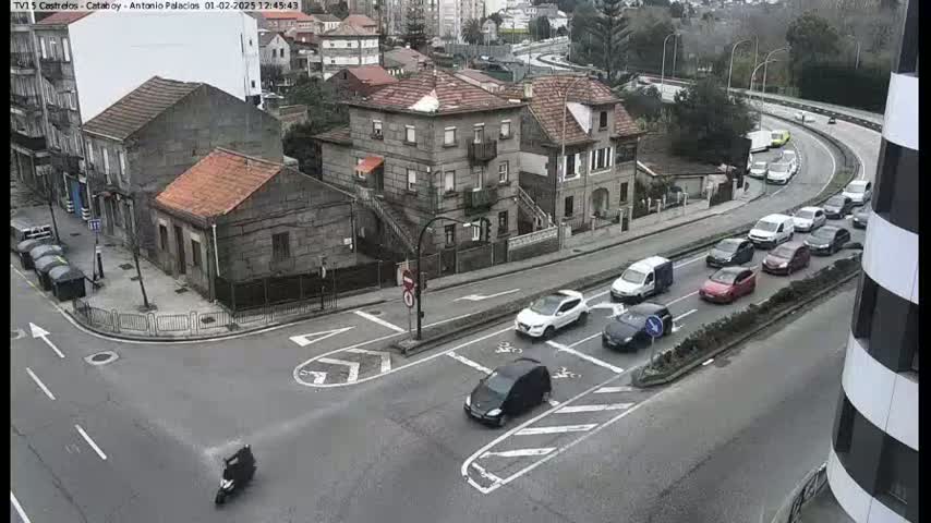 A street intersection with several cars and a scooter, surrounded by buildings on an overcast day.