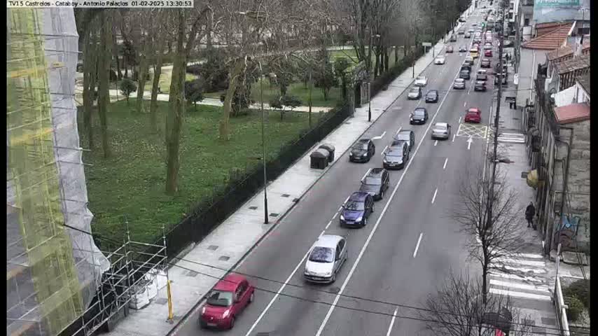 A road with moderate traffic flows next to a park and a building under construction, on a partly cloudy day.