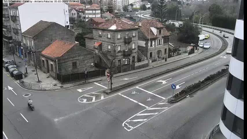 A high-angle view of a street intersection with several buildings, a curved road with several cars, and a person on a motorcycle, under overcast conditions.