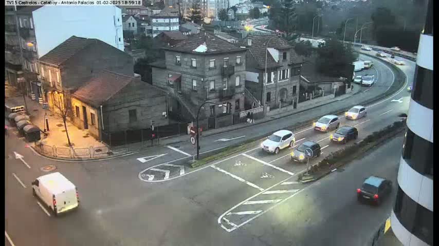 A high-angle, twilight view of a street intersection with several cars and a van, showing some buildings and a curved road in the background under overcast skies.