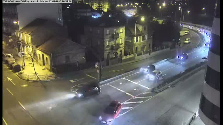 A nighttime, high-angle view of a city intersection with several cars driving through, some buildings and a curved road visible in the background.