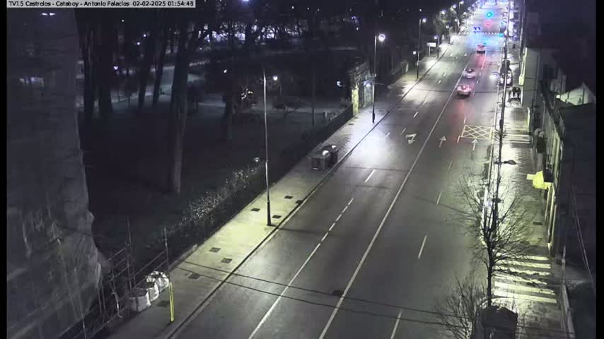 A nighttime, high-angle view shows a mostly empty, brightly lit road with a few cars driving, flanked by a park on one side and buildings on the other.