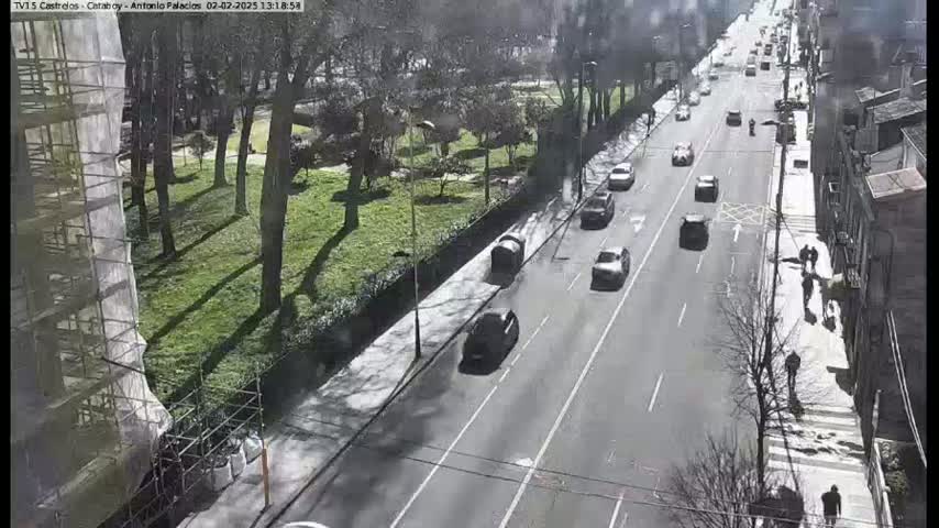 A sunny day reveals a city street with cars driving on it, pedestrians walking on the sidewalk next to a park with trees and a building under construction.