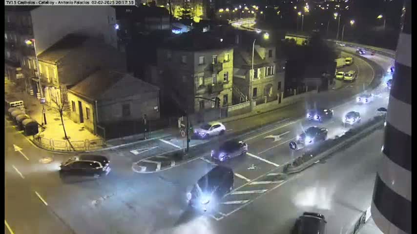 A high-angle nighttime view shows several cars driving on wet roads around a city intersection near buildings.