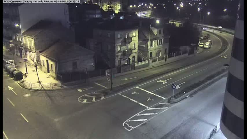 An aerial nighttime view shows a street intersection with several buildings, some cars driving on a curving road, and streetlights illuminating the scene.