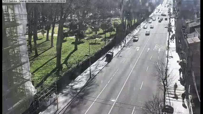 A sunny day shows a partly-snowy city street with cars driving on a multi-lane road that passes a park with leafless trees and a building under construction.