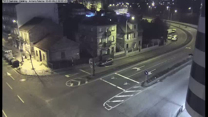 A nighttime aerial view of a street intersection with several buildings and a few cars, illuminated by streetlights.