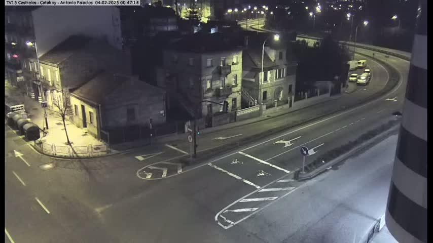 A nighttime aerial view shows a street intersection with several buildings, and a highway curving away in the background with a few vehicles.