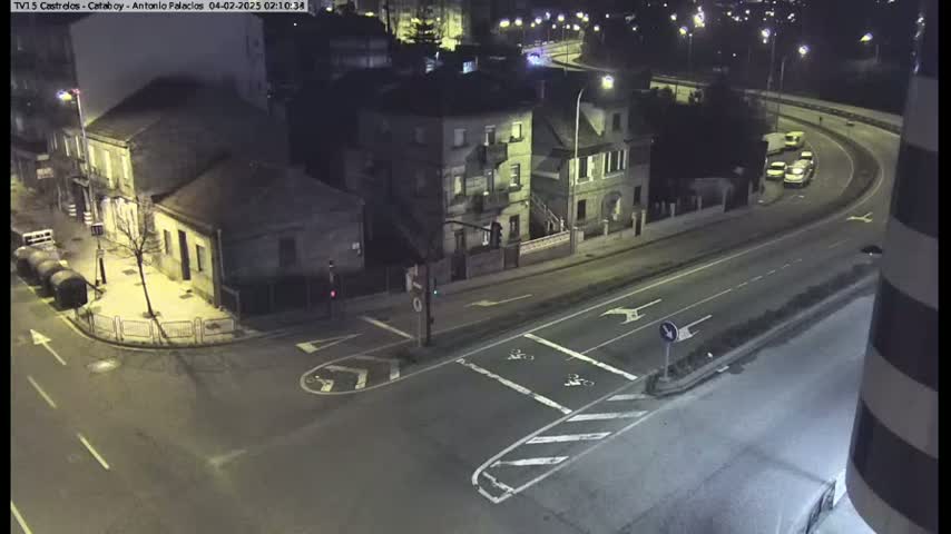 A nighttime aerial view shows a street intersection with several buildings and a few vehicles on a curving road under artificial street lighting.