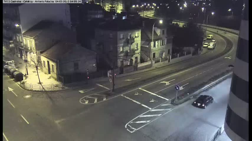 A nighttime, high-angle view shows a street intersection with several buildings and a few cars driving on the roads.