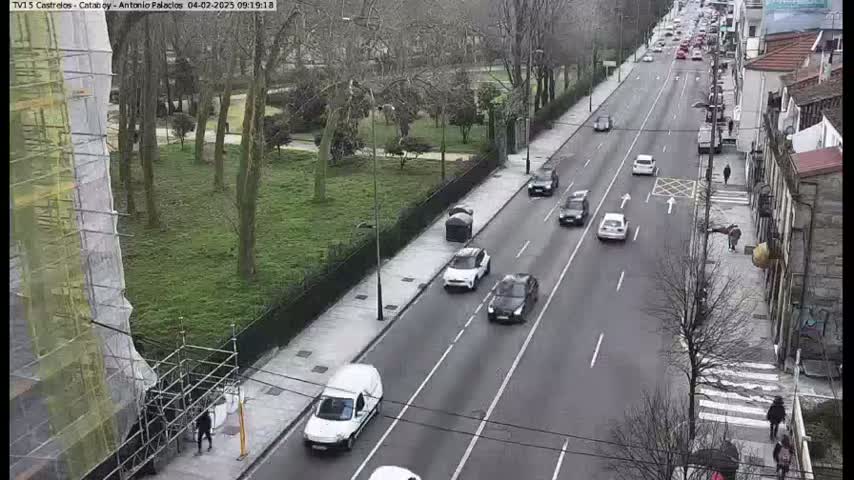 A street scene shows several cars driving on a multi-lane road that borders a park with leafless trees under an overcast sky.
