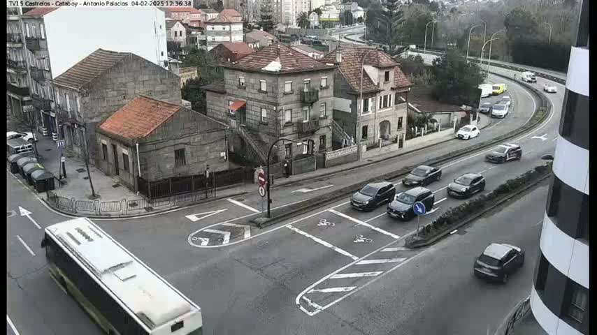 A long white bus is driving past a multi-lane intersection with several cars and stone buildings, under an overcast sky.