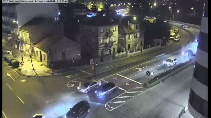 A nighttime aerial view of a street intersection with several cars driving through, under partly cloudy conditions.