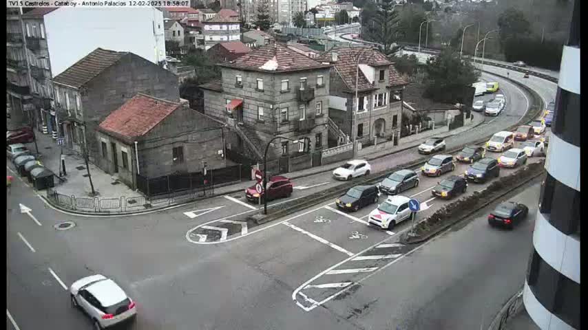 A street intersection in an overcast city, surrounded by stone buildings, with numerous cars driving on the roads and parked along the curb.