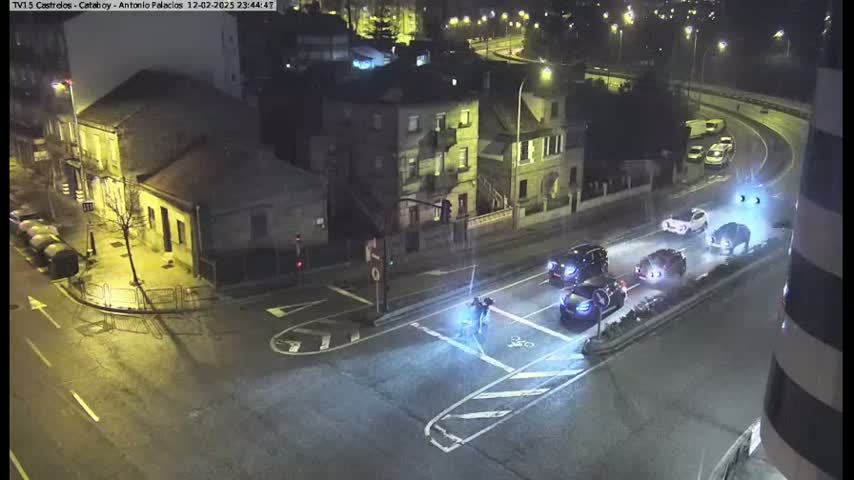 At night, under wet conditions, several cars are driving through a street intersection where two pedestrians are crossing.