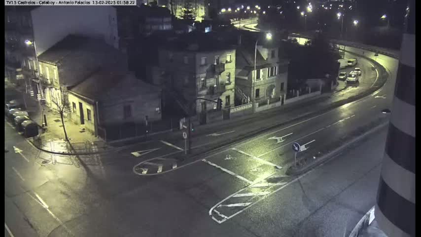 A wet, nighttime street intersection shows several buildings, parked cars, and a few vehicles on the road curving away from the camera.