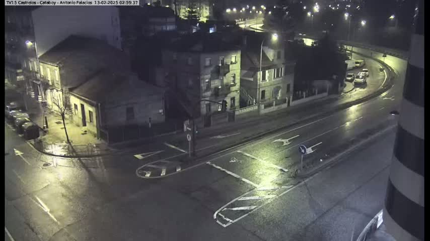 A rain-slicked street intersection at night, showing several buildings and a few vehicles on the road.