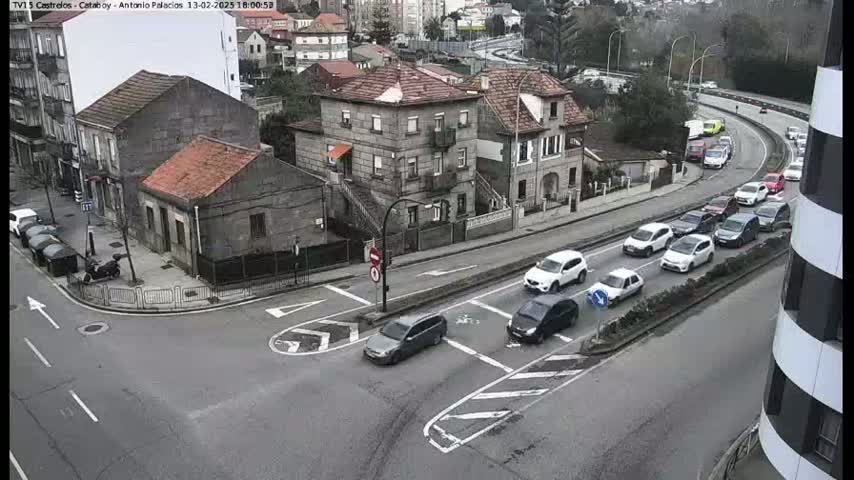 A high-angle view shows a street intersection with several cars at a standstill and moving, along with several buildings, under an overcast sky.