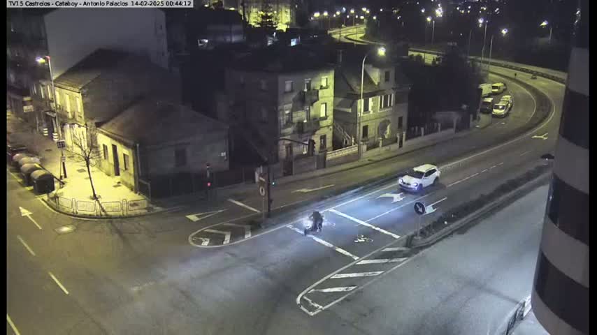 A nighttime overhead view of a street intersection with several buildings, a few cars, and a person on a scooter or motorcycle crossing the street under bright streetlights.