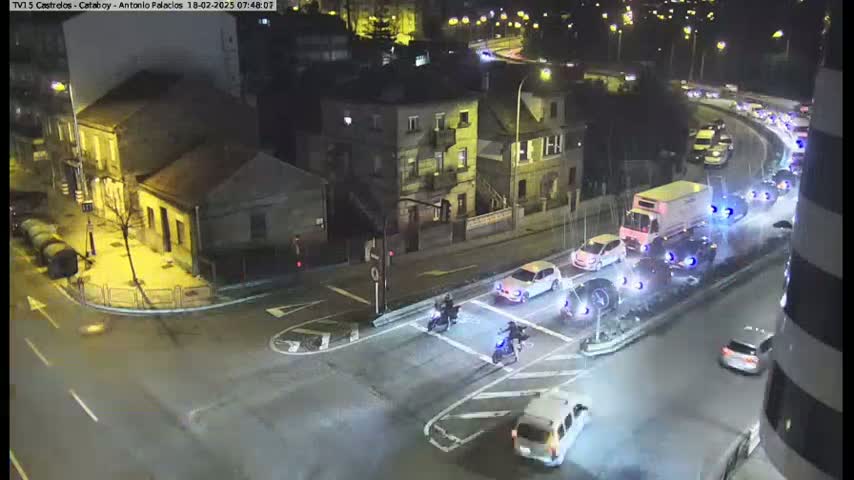At night, several vehicles are seen driving on city roads and intersecting at a crosswalk where motorcyclists are also present.