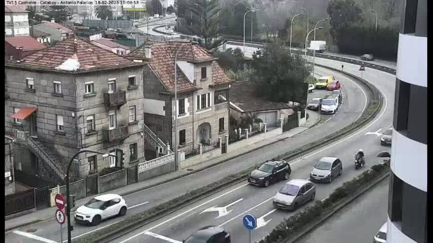 A high-angle view shows several cars driving on a curved road in front of a stone building, with overcast skies.