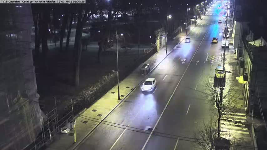 A nighttime view of a mostly empty road with a few cars driving and parked along the sides, lined with buildings and trees.
