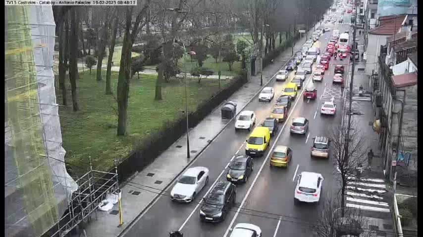 A wet city street scene shows a long line of slow-moving traffic, alongside a park with leafless trees, and a building under construction.