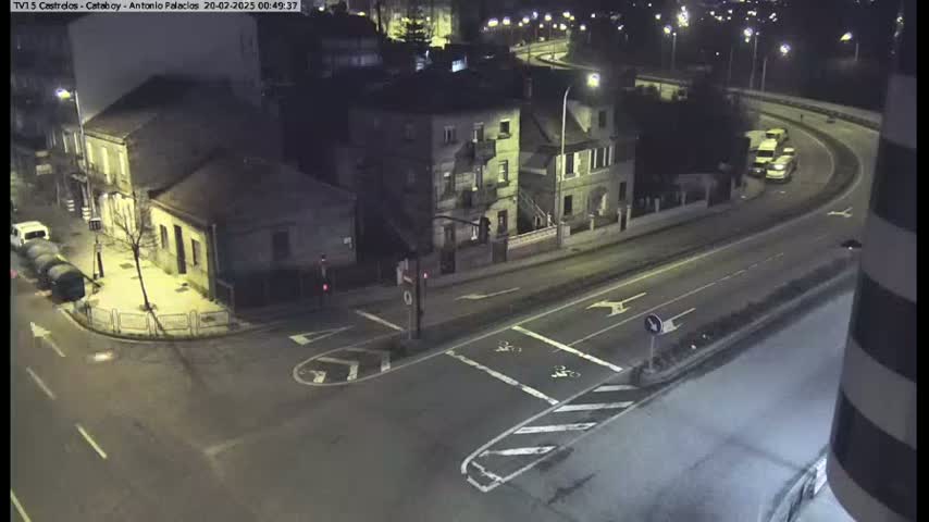 A nighttime aerial view shows a street intersection with several buildings and a few vehicles on a curving road.