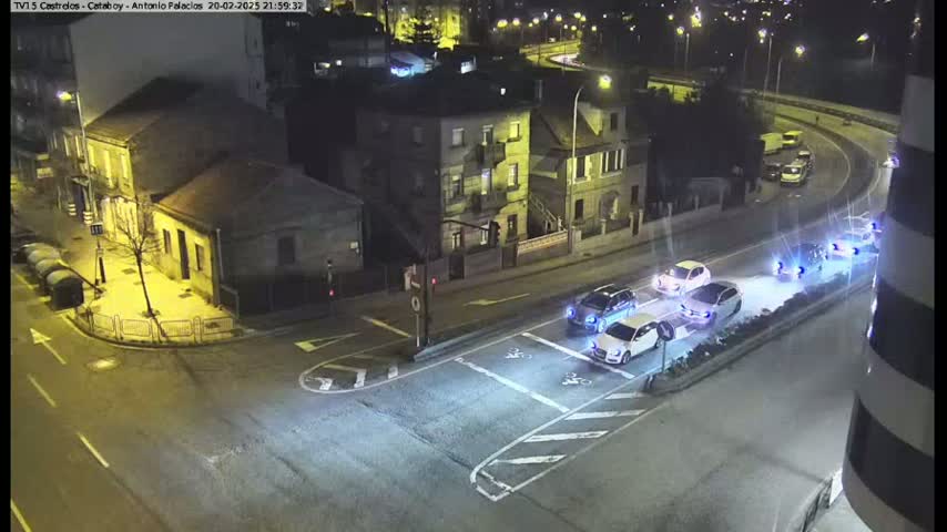 At night, several cars are stopped at a street intersection in front of buildings under bright streetlights.