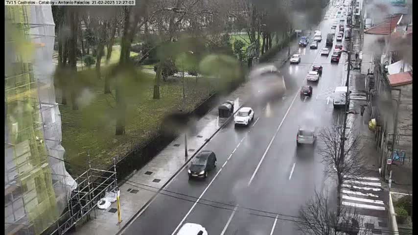 A rainy day scene shows a wet roadway with several cars driving, alongside a park and a building under construction.