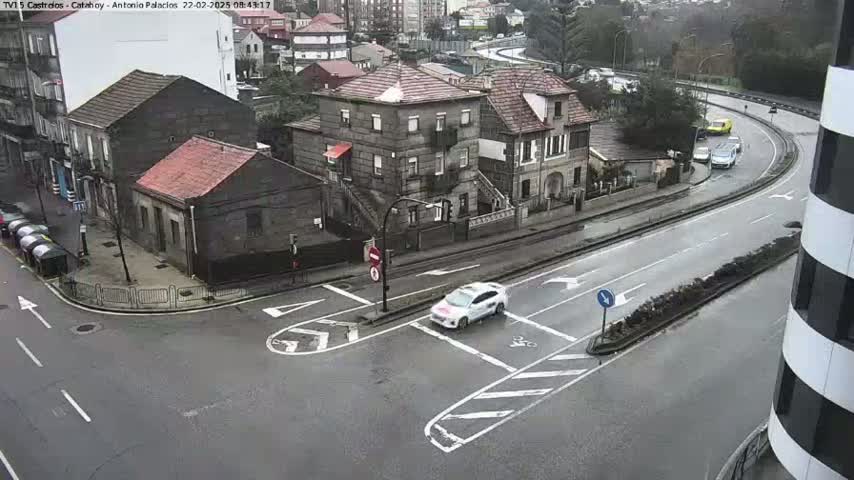 A wet street intersection with several buildings and a few cars, one of which is white, is seen from an elevated viewpoint on an overcast day.